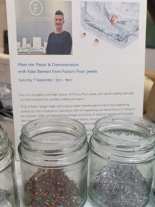 A poster for Meet The Maker - Factory Floor Jewels at the Bluecoat Display Centre with some pots of bronze and aluminium swarf infront of them