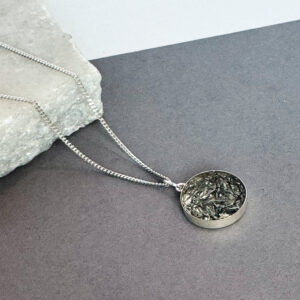 photo of a handmade sterling silver pendant containing flecks of aluminium set in resin. It is shown on a grey background with the chain on a white piece of marble