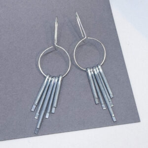 a pair of silver and steel earrings on a grey background. They are inspired by art deco design and feature a circle with 5 steel pins hanging from it.