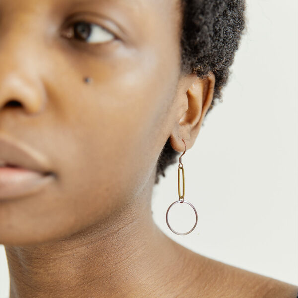 A black woman with short hair is wearing a brass and steel earring by Factory Floor Jewels - only one ear is visible